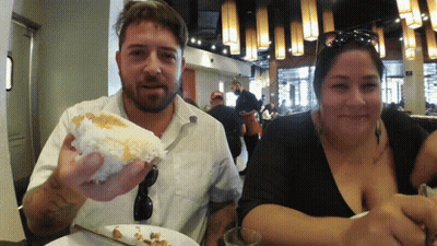 A Man And A Woman Are Sitting At A Table Eating Food GIF
