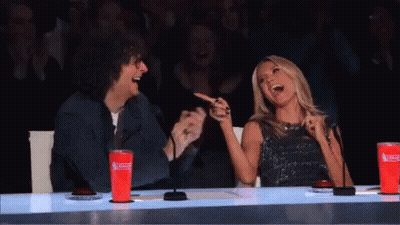A Man And A Woman Are Sitting At A Table Laughing . The Woman Is Pointing At The Man . GIF