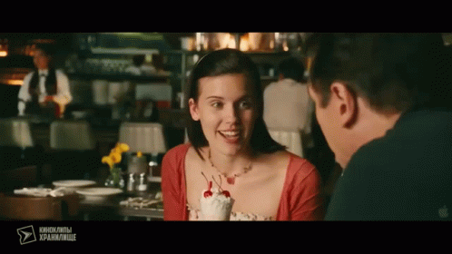 A Man And A Woman Are Sitting At A Table With A Cherry On Top Of An Ice Cream Sundae GIF