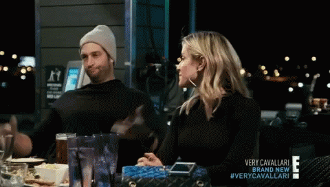 A Man And A Woman Are Sitting At A Table With A Sign That Says Very Cavallari On It GIF