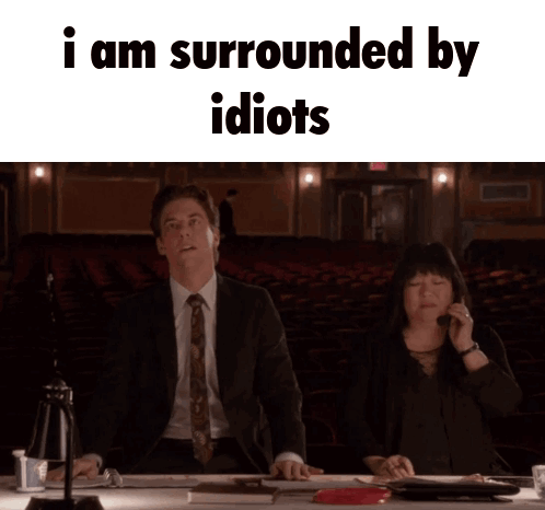 A Man And A Woman Are Sitting At A Table With The Words I Am Surrounded By Idiots GIF
