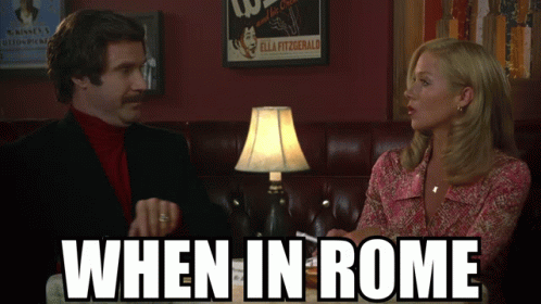 A Man And A Woman Are Sitting At A Table With The Words When In Rome Below Them GIF