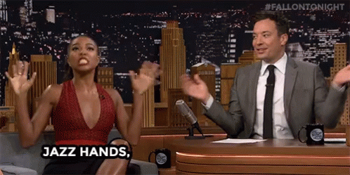 A Man And A Woman Are Sitting At A Table With Their Hands In The Air . The Woman Is Saying Jazz Hands . GIF