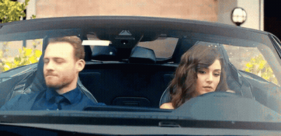 A Man And A Woman Are Sitting In A Car Looking Out The Window GIF