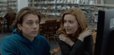 A Man And A Woman Are Sitting In A Library Looking At A Computer Screen GIF