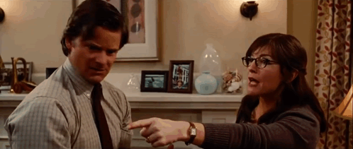 A Man And A Woman Are Sitting In A Living Room And The Woman Is Pointing At The Man 'S Shirt . GIF