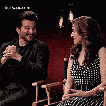 A Man And A Woman Are Sitting In Chairs With Their Hands Folded . GIF