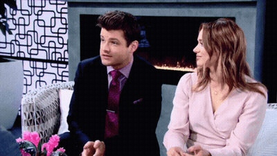 A Man And A Woman Are Sitting In Front Of A Fireplace GIF