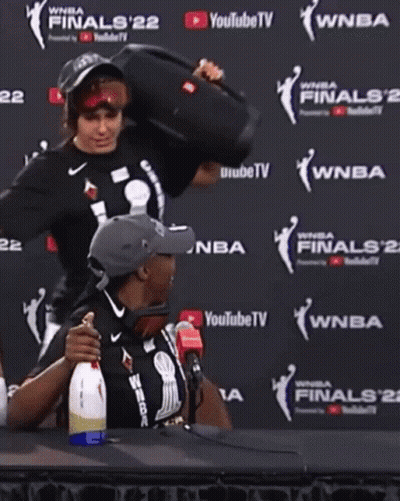 A Man And A Woman Are Sitting In Front Of A Wall That Says Wnba Finals 22 GIF
