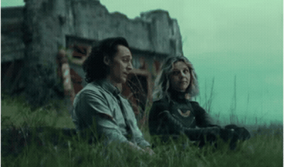 A Man And A Woman Are Sitting In The Grass Next To A Building . GIF