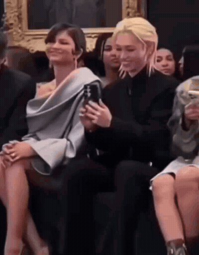 A Man And A Woman Are Sitting Next To Each Other At A Fashion Show . The Woman Is Holding A Cell Phone . GIF
