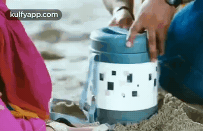A Man And A Woman Are Sitting On A Beach Holding A Thermos . GIF