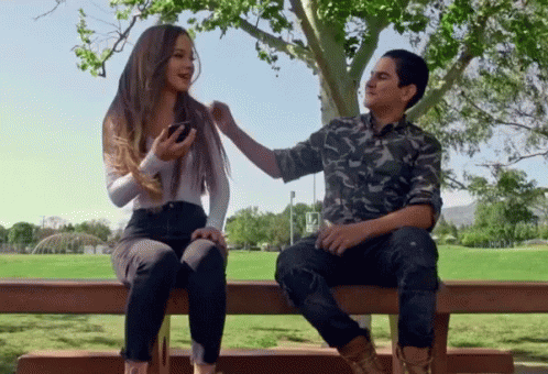 A Man And A Woman Are Sitting On A Bench Under A Tree And The Man Is Touching The Woman 'S Hair GIF
