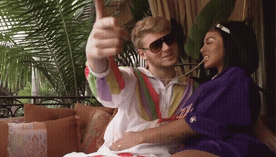 A Man And A Woman Are Sitting On A Couch And The Man Is Wearing Sunglasses And Giving A Thumbs Up GIF