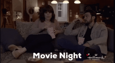A Man And A Woman Are Sitting On A Couch Eating Popcorn And Watching A Hallmark Movie GIF