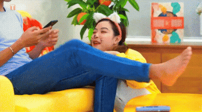 A Man And A Woman Are Sitting On A Couch Looking At Their Phones . The Woman Is Wearing Cat Ears . GIF