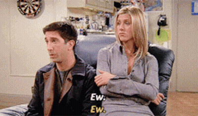 A Man And A Woman Are Sitting On A Couch With A Dart Board In The Background GIF