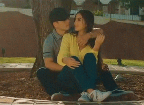 A Man And A Woman Are Sitting Under A Tree In A Park . GIF