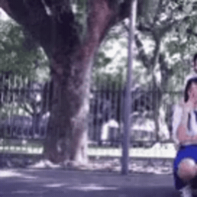 A Man And A Woman Are Sitting Under A Tree On The Side Of The Road . GIF