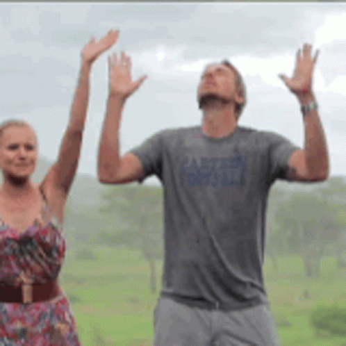 A Man And A Woman Are Standing In A Field With Their Hands Up GIF