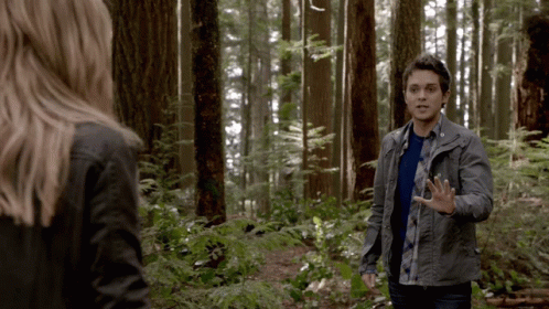 A Man And A Woman Are Standing In A Forest Talking To Each Other GIF