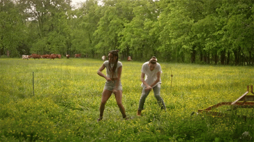 A Man And A Woman Are Standing In A Grassy Field With Cows In The Background GIF