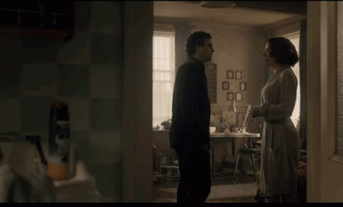 A Man And A Woman Are Standing In A Room With Pictures On The Wall And A Table GIF