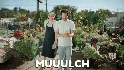 A Man And A Woman Are Standing In Front Of A Bunch Of Potted Plants And The Word Muuulch Is Visible GIF