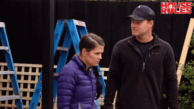 A Man And A Woman Are Standing In Front Of A Fence That Says House Rules GIF