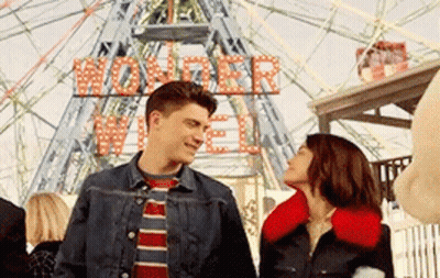 A Man And A Woman Are Standing In Front Of A Ferris Wheel That Says Wonder Wheel On It GIF