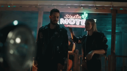 A Man And A Woman Are Standing In Front Of A Motel Sign GIF