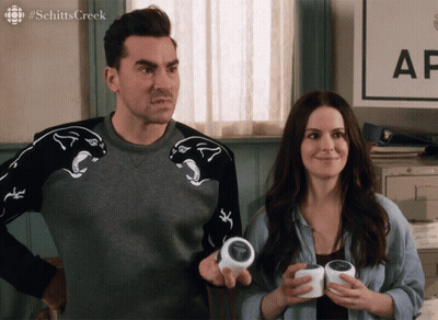 A Man And A Woman Are Standing In Front Of A Sign That Says Schitts Creek On It GIF