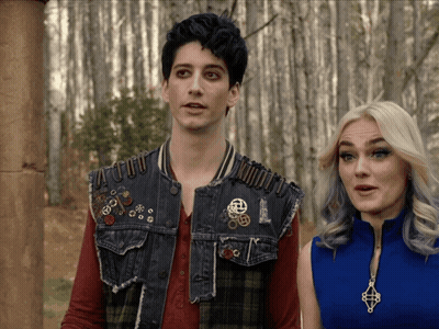 A Man And A Woman Are Standing Next To Each Other And The Man Is Wearing A Vest With Pins On It GIF