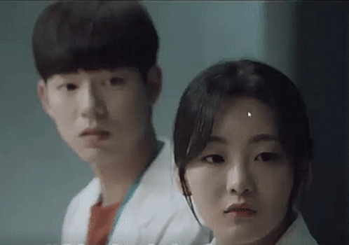 A Man And A Woman Are Standing Next To Each Other In A Room And Looking At The Camera . GIF