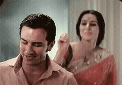 A Man And A Woman Are Standing Next To Each Other In A Room . The Woman Is Holding The Man 'S Hair . GIF