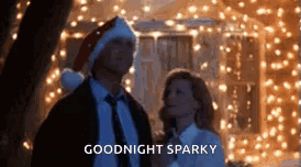 A Man And A Woman Are Standing Next To Each Other In Front Of A Christmas Tree . GIF