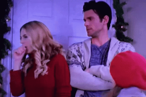 A Man And A Woman Are Standing Next To Each Other In Front Of A Christmas Tree . The Woman Is Coughing . GIF