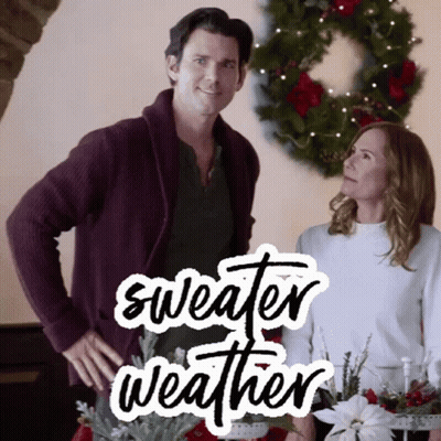 A Man And A Woman Are Standing Next To Each Other In Front Of A Christmas Wreath With The Words Sweater Weather Written  GIF