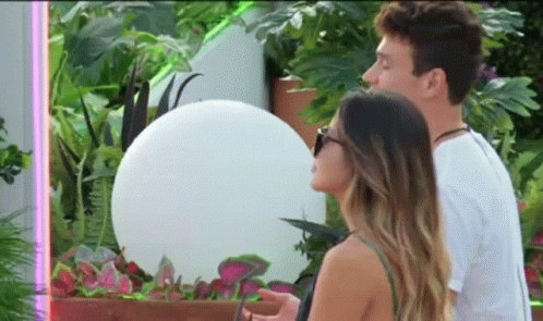 A Man And A Woman Are Standing Next To Each Other In Front Of A Plant . GIF