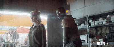 A Man And A Woman Are Standing Next To Each Other In Front Of A Shelf With Netflix Written On It GIF