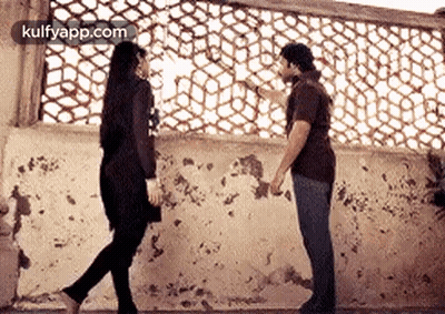 A Man And A Woman Are Standing Next To Each Other In Front Of A Wall Holding Hands . GIF