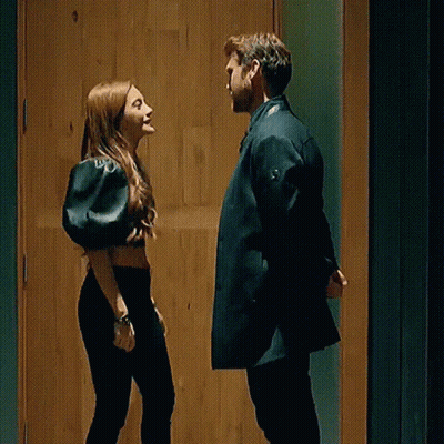 A Man And A Woman Are Standing Next To Each Other In Front Of A Wooden Door And Looking At Each Other . GIF