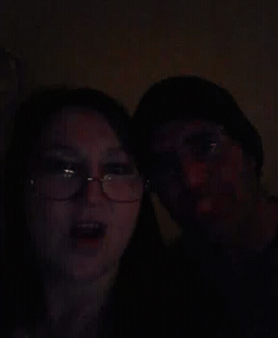 A Man And A Woman Are Standing Next To Each Other In The Dark . The Woman Is Wearing Glasses And A Hat . GIF