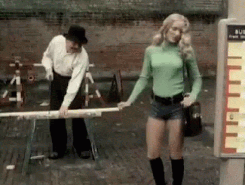 A Man And A Woman Are Standing Next To Each Other On A Sidewalk . The Woman Is Wearing Shorts . GIF