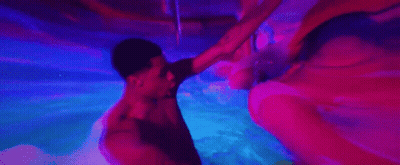 A Man And A Woman Are Swimming Underwater In A Pool With Purple Lights . GIF