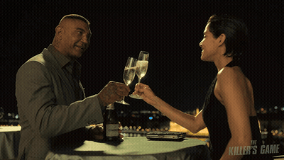 A Man And A Woman Are Toasting With Champagne And The Killer 'S Game Is Visible In The Background GIF