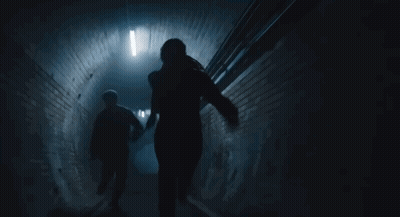 A Man And A Woman Are Walking Down A Dark Tunnel Holding Hands . GIF