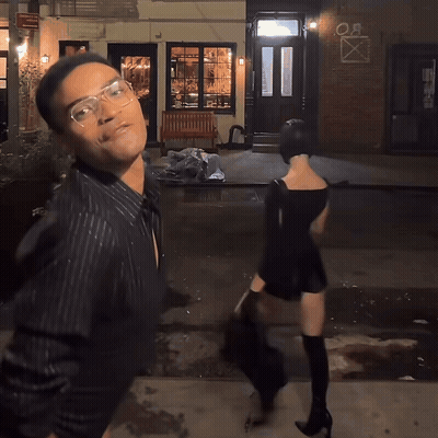 A Man And A Woman Are Walking Down A Street In Front Of A Building That Says " Oa " On It GIF