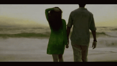 A Man And A Woman Are Walking On The Beach Holding Hands . The Woman Is Wearing A Green Dress . GIF