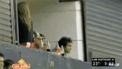 A Man And A Woman Are Watching A Game With A Sign That Says Las Noticias On It GIF